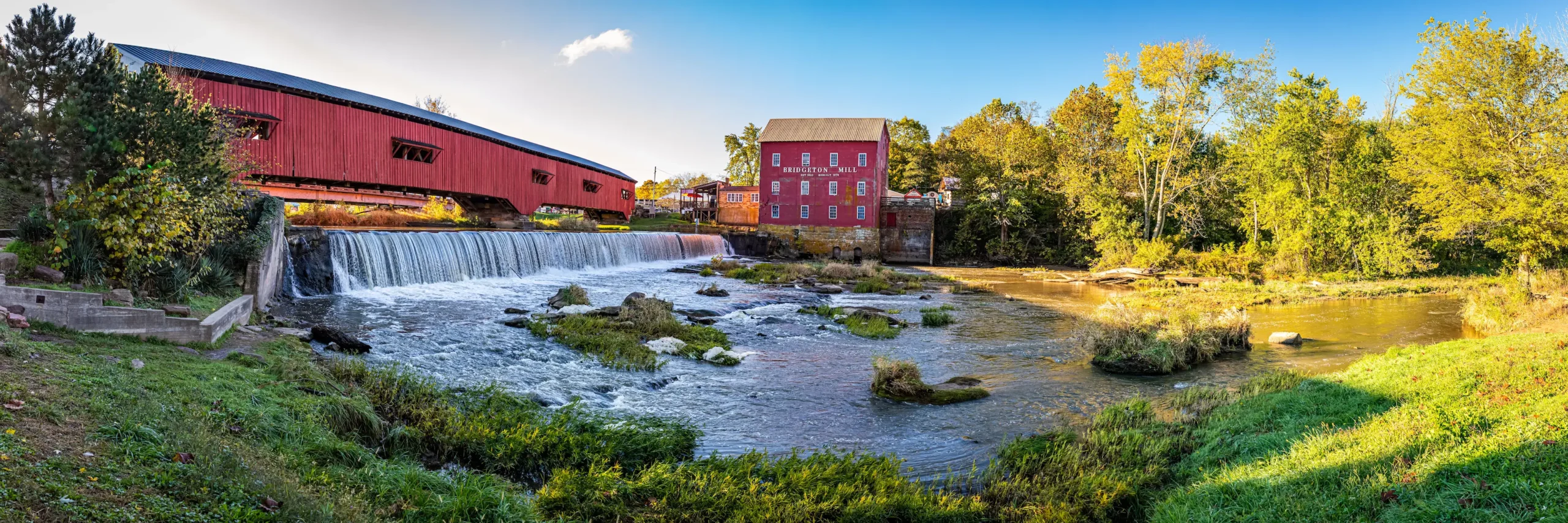 Scenic view of the Bridgeton Covered Bridge and historic Bridgeton Mill in Parke County, Indiana, with a small waterfall and lush green trees under a clear blue sky.